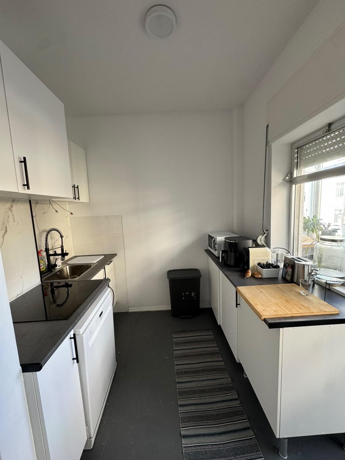 Cozinha com móveis brancos de puxadores pretos, bancada em tom grafite, placa de indução, lava-loiça em inox e revestimento em efeito mármore claro.